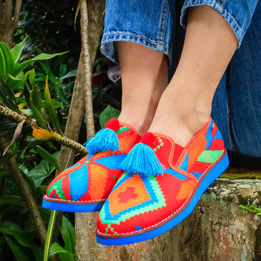 "Mocasines artesanales Walekeru en tonos rojos, azules y verdes con borlas tejidas, inspirados en la tradición colombiana."